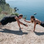 A couple performs a workout on a cliff overlooking the sea, embracing outdoor fitness and healthy living.