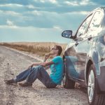 Man sitting by a car on a remote gravel road under a cloudy sky, deep in thought.